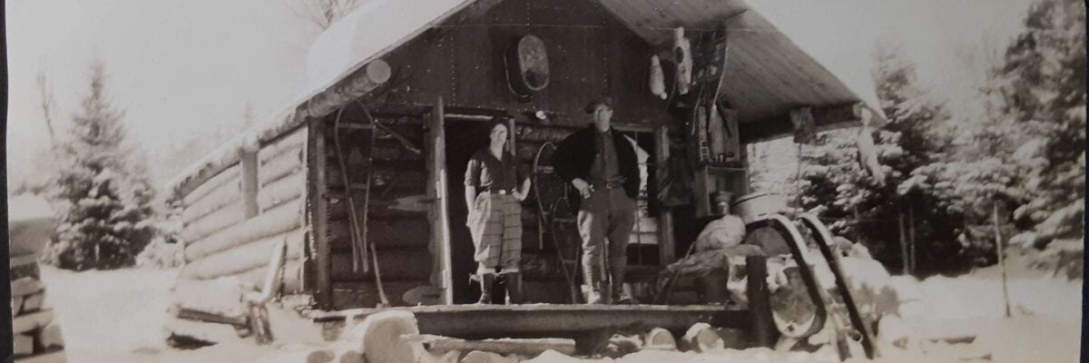 black and white photo of two men standing in front of cabin in winter