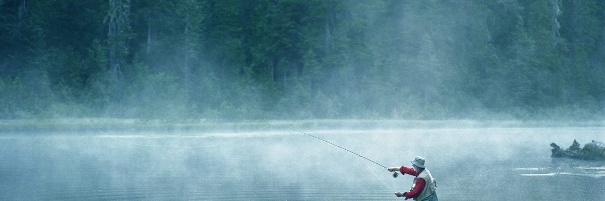 Senior fly fisherman standing in lake covered with fog, casting line